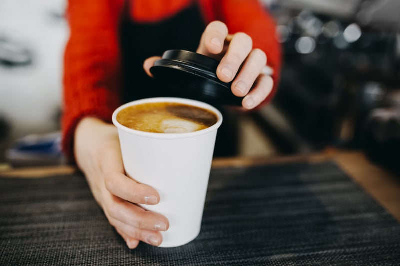 A person in a red sweater and black apron hands over a white take-out cup of latte with a black lid.