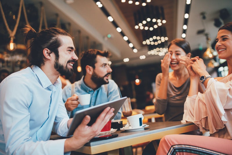Four friends, two men and two women, happily chatting and laughing at a cafe. The man in front holds a tablet.