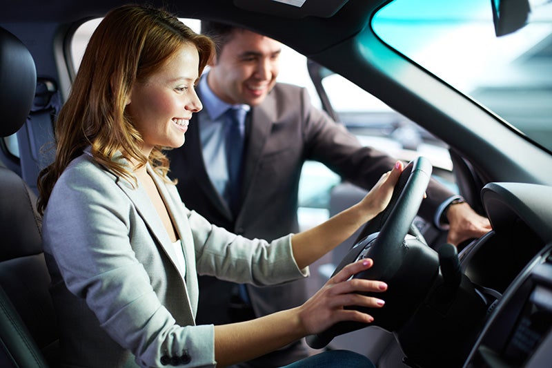 A smiling woman in a light jacket sits in the driver's seat of a car, a salesperson smiling beside her.