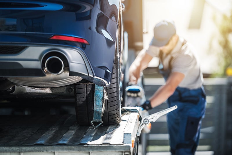 A blue car is secured to a tow truck bed by a strap around its rear wheel, with a worker in the background.
