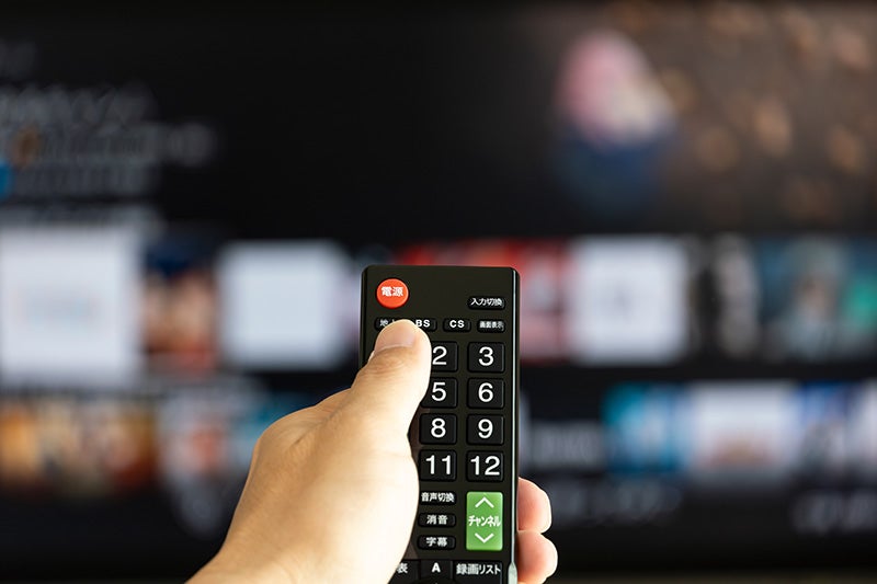 A hand presses a button on a black TV remote control, with a blurred TV screen in the background.