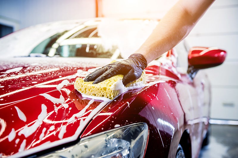A person wearing a black glove uses a yellow sponge to wash a red car covered in white soap suds.